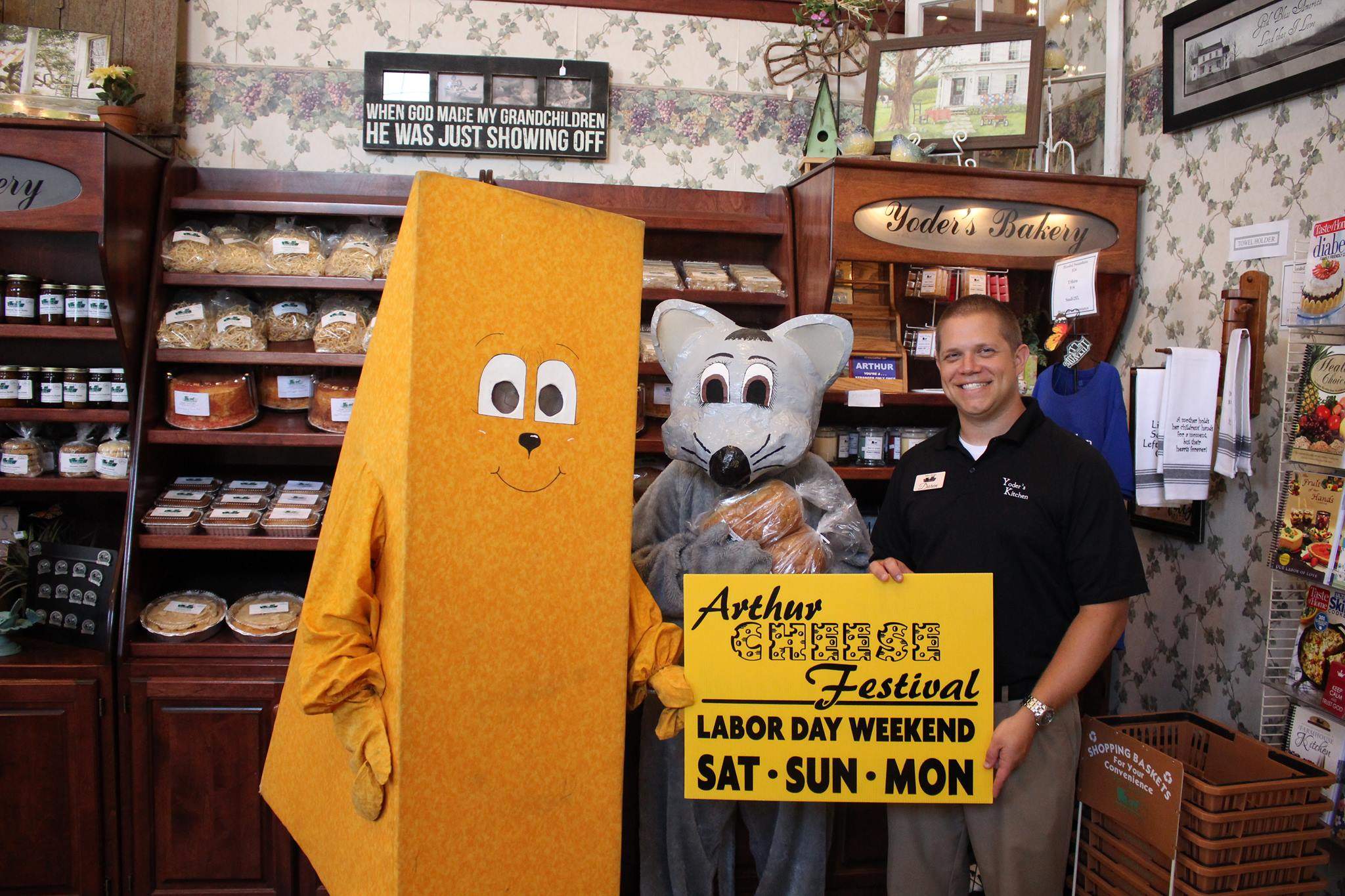 Maskottchen in einem Käsekostüm und einem Mäusekostüm stehen neben einem Mann, der ein Schild des Arthur Cheese Festival hält