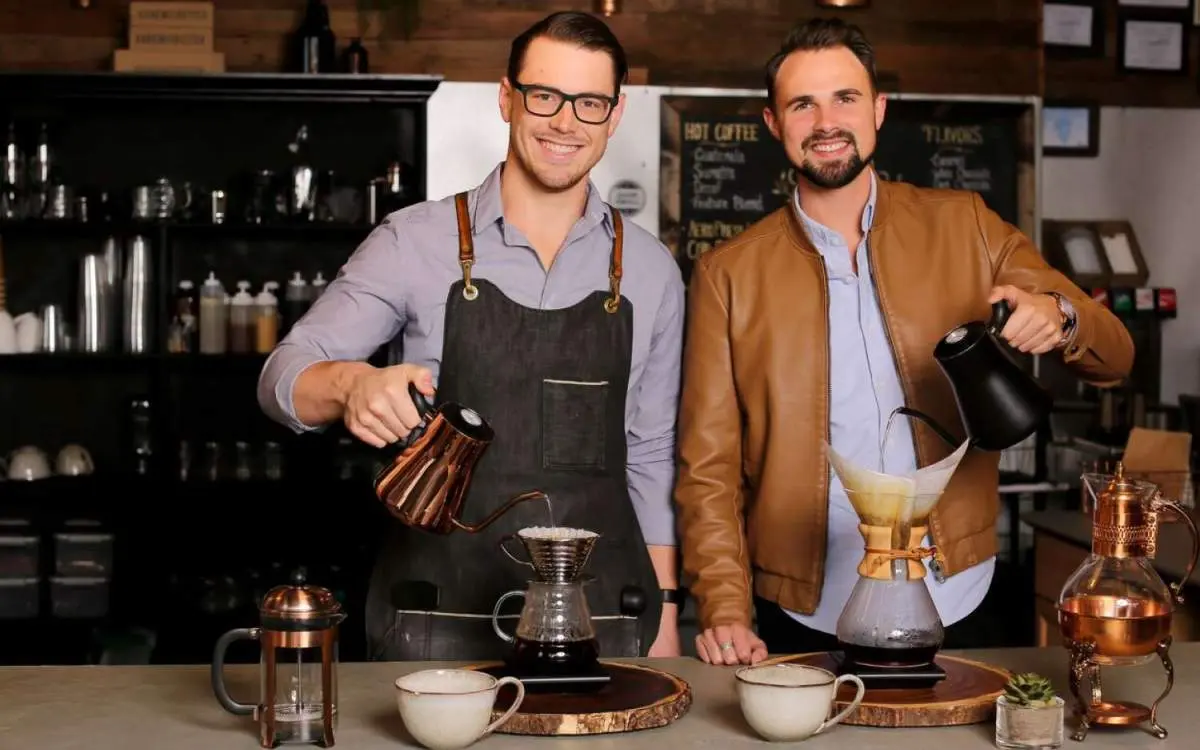 Zwei Männer schauen in die Kamera, während sie in einem Café Filterkaffee einschenken