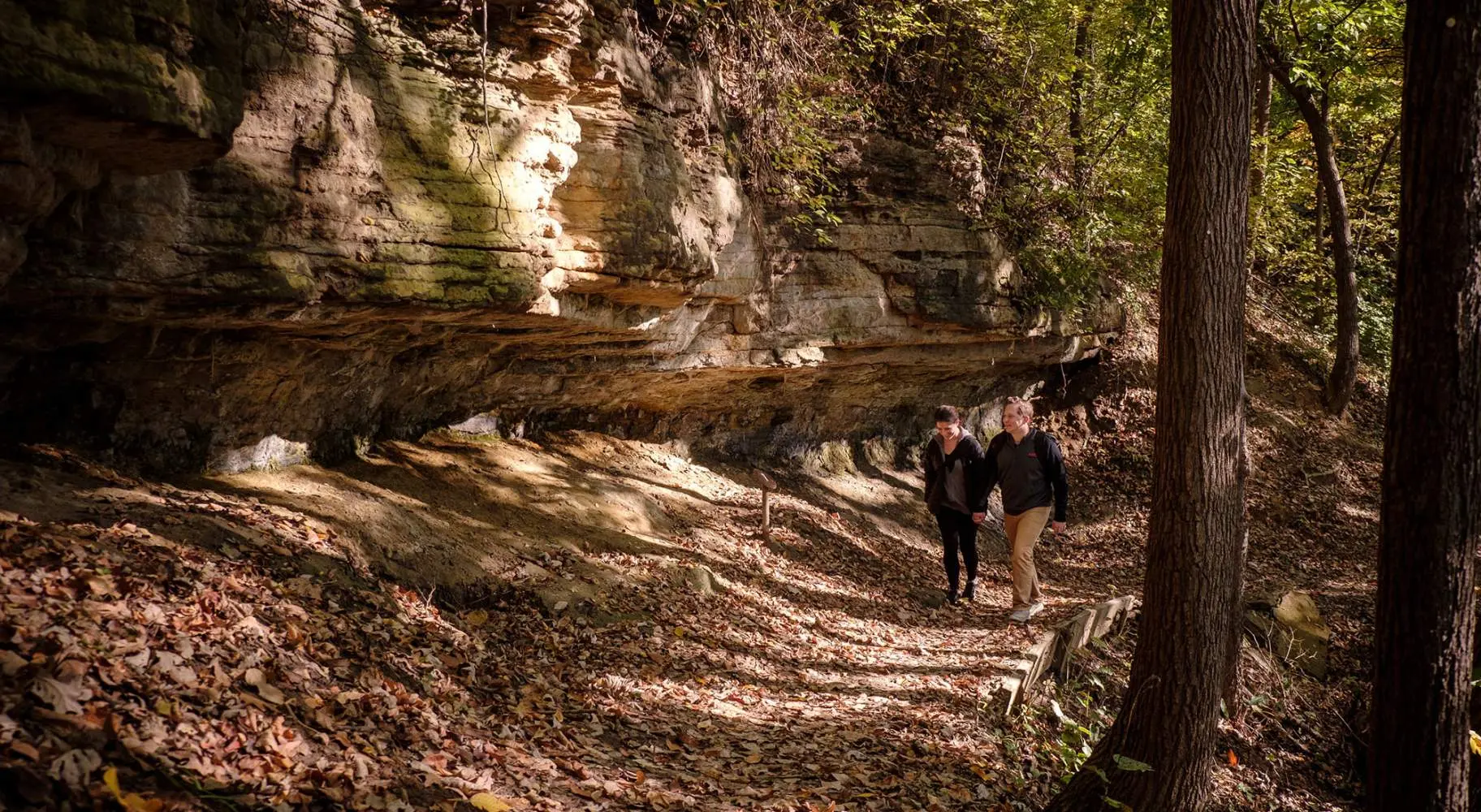 Zwei Menschen gehen im Wald entlang eines großen Felsens