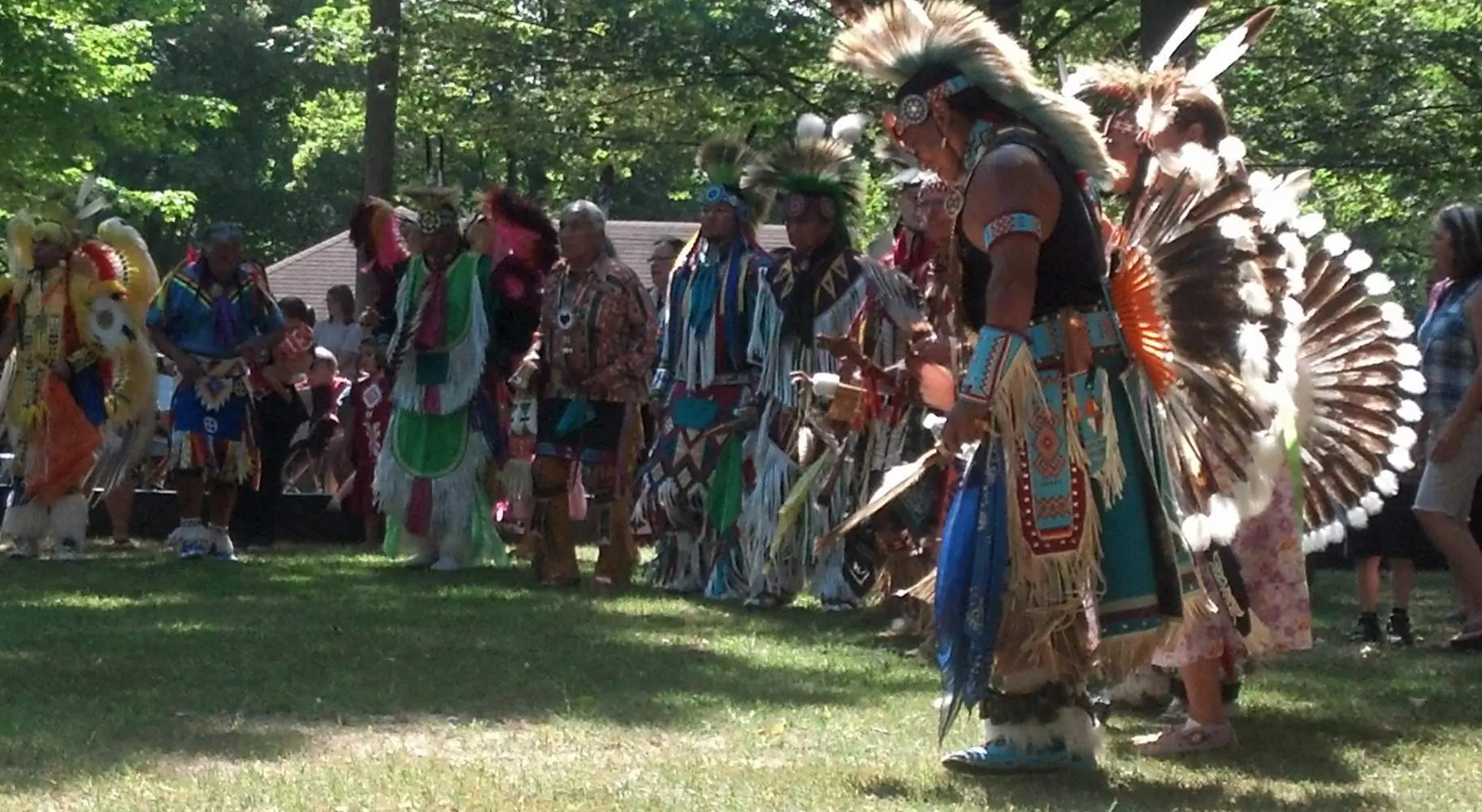 Eine Gruppe von Männern in indianischer Kleidung auf einer Graslichtung inmitten von Bäumen