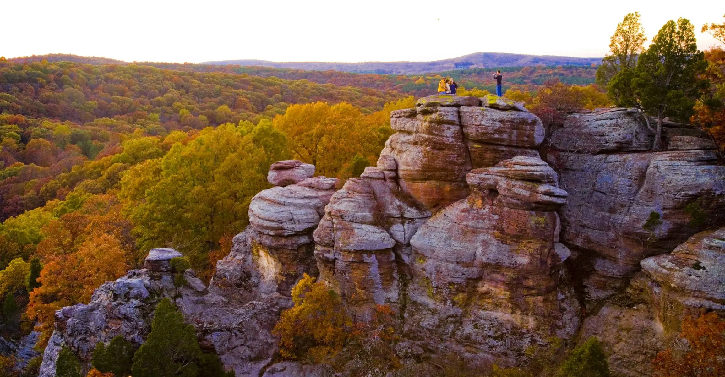 Eine entfernte Gruppe von vier Personen posiert für ein Foto auf einer felsigen Klippe mit Blick auf Wälder aus grünen und goldenen Bäumen