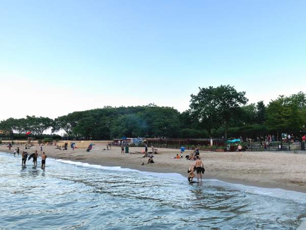 Kleine Gruppen von Menschen an einem ruhigen Strand am Seeufer in der Abenddämmerung, mit niedrigen grünen Bäumen im Hintergrund