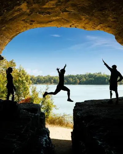 Eine Person springt über einen Felsen mit Freunden, blauem Himmel und Wasser im Hintergrund