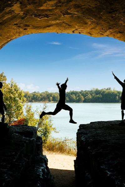 Eine Person springt über einen Felsen mit Freunden, blauem Himmel und Wasser im Hintergrund