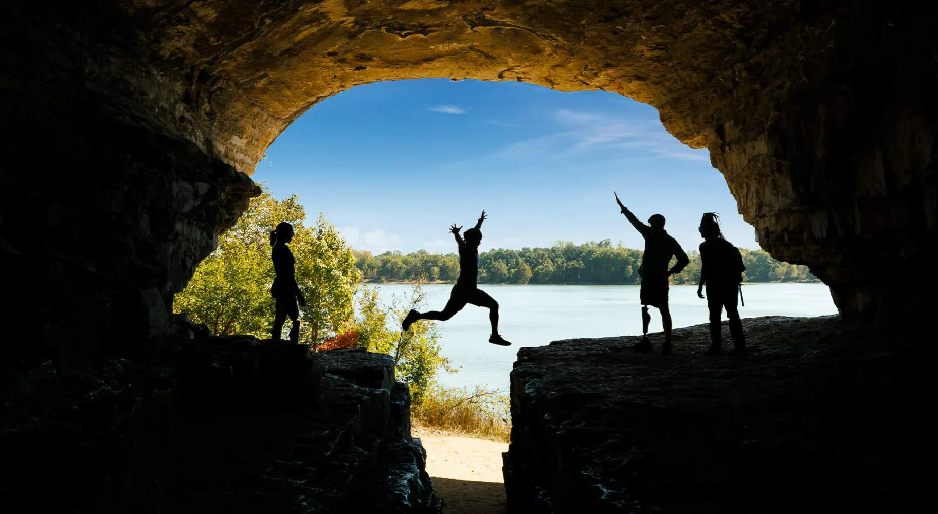 Eine Person springt über einen Felsen mit Freunden, blauem Himmel und Wasser im Hintergrund