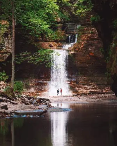 Zwei Menschen stehen vor einem Wasserfall, neben einem ruhigen Teich.