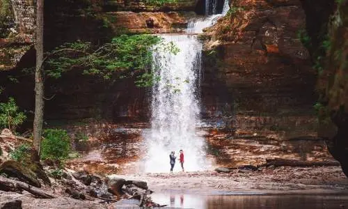 Zwei Menschen stehen vor einem Wasserfall, neben einem ruhigen Teich.
