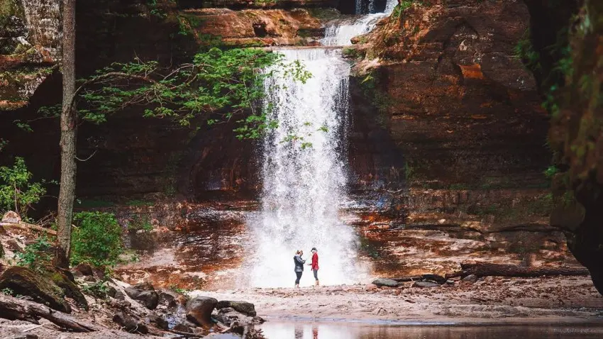 Zwei Menschen stehen vor einem Wasserfall, neben einem ruhigen Teich.