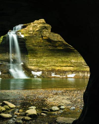 Von einer Höhle aus gesehen stürzt ein Wasserfall in ein Becken, das von schneebedeckten Felswänden umgeben ist.