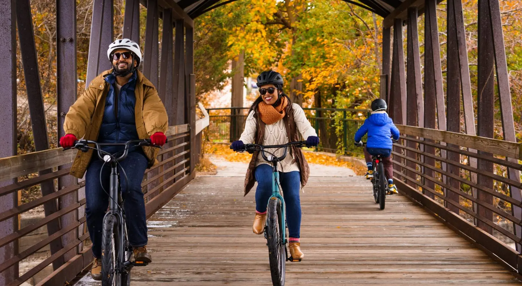Ein Paar fährt mit dem Fahrrad auf einer alten überdachten Holzbrücke, mit Herbstlaub im Hintergrund