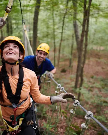 Zwei Personen, die Helme und Klettergurte tragen, bewältigen einen Hochseilgarten im Wald.