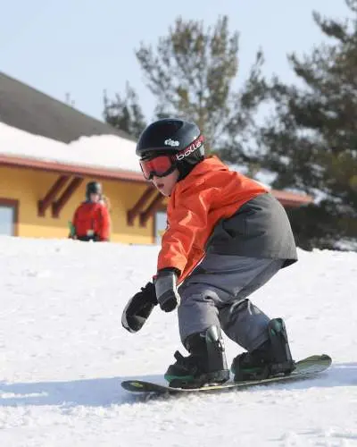 Ein kleines Kind fährt auf einem Snowboard durch den Schnee, trägt einen Helm, eine Brille und warme Skikleidung