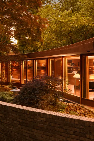Ein modernes Haus aus gebogenem Glas, das in der Abenddämmerung beleuchtet ist und von Bäumen und einer niedrigen Ziegelmauer umgeben ist.
