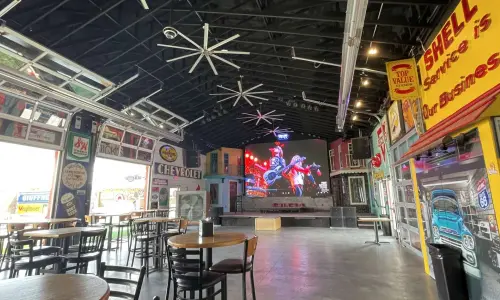 Innenansicht einer geräumigen Bar und Live-Musik-Location mit Tischen, Vintage-Autoschildern und -Dekoration sowie einer großen Leinwand über der Bühne.